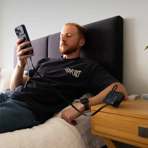 Man lying on a bed using a smartphone with a power bank attached, in a bedroom setting.
